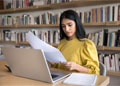 A woman sits at a desk, with a laptop in front of her. She is holding a document and considering the information in it.