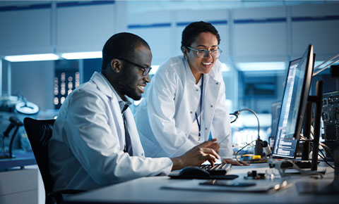 A man sits at a computer desk and a woman stands near by. Both look at the computer screen