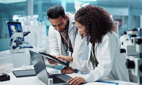 Two people wearing lab coats, look at information on a tablet, while standing in a lab. There is a microscope on the table near them.