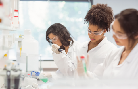 Three students work in a lab. Each of them wears a lab coat and protective goggles.