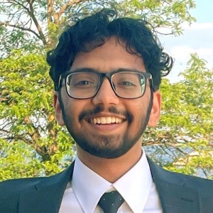 Headshot of a young man with dark, curly hair and glasses, wearing a suit.