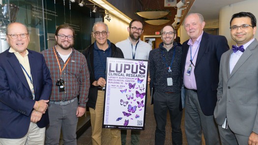 Group of men standing behind a Lupus poster.